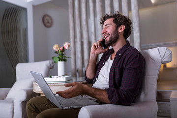 Fototapeta premium Young attractive smiling guy is browsing at his laptop and talking oh mobile phone, sitting at home on the cozy sofa, wearing casual outfit. Freelance business work from home concept