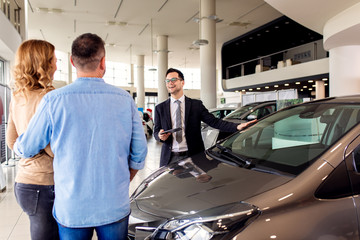 Young sales agent helping adult couple to choose a new car in modern car showroom.