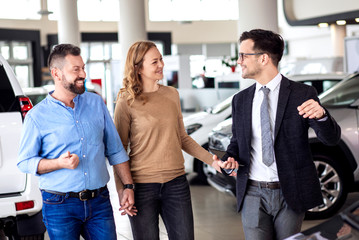 Young sales agent helping adult couple to choose a new car in modern car showroom.