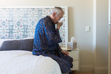 Elderly man sitting on bed blowing nose - coronavirus concept (selective focus)