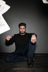 A young dark-haired man with a beard sits on the floor and throws papers and documents into the air. Human emotions: rage, anger, discontent. Close-up studio portrait of a man.