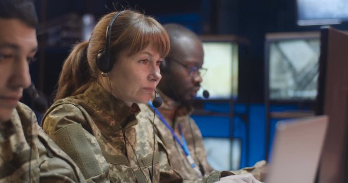 Mixed-races male and female militarians in camouflage uniforms and headsets working at computers in monitoring operation office. Close up of officers dispatchers passing information to army.