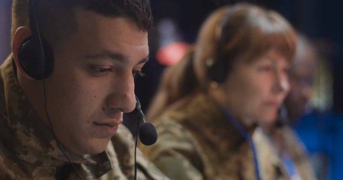 Close up of Caucasian male soldier in headset working as dispatcher in conflict zone controlling office. Blurred woman and African Ameican man colleagues on background.