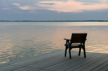 Fototapeta na wymiar Wooden chair on a wood pier overlooking calm lake at sunset skyline.