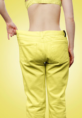 A Caucasian woman shows the results of her weight loss by pulling off wide-waisted yellow jeans. Isolate, yellow background. Rear view. The concept of weight loss, sports and diet