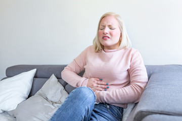 Fototapeta premium View of young woman suffering from stomachache on sofa at home. Woman sitting on bed and having stomach ache. Young woman suffering from abdominal pain while sitting on sofa at home