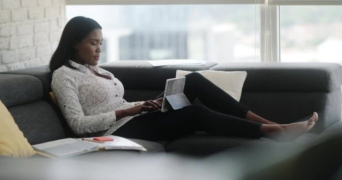 Black woman working from home during quarantine. African american people and mobile work, doing telework. Young businesswoman at work with laptop computer, lying on sofa