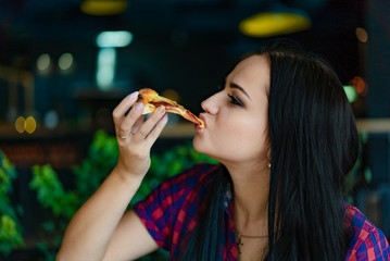 Beautiful brunette girl in t-shirt eating pizza at restaurant. A pretty girl feels happy and enjoys eating a piece of delicious pizza.