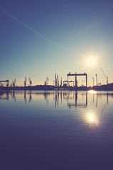 Shipyard cranes silhouettes reflected in water at sunset, color toning applied. - obrazy, fototapety, plakaty