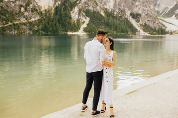 Happy moments together. Happy young couple embracing and smiling while standing near the lake Braies, Italy. Couple cuddling affectionate on the beach in summer with the lake in the background.