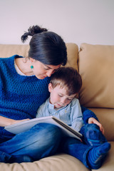 Sick little boy listening to a story from a book. Lifestyle with children at the house. Little kid grounded at home. Young mother reading the homework to her child with no school time.