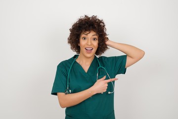 Joyful pretty young doctor woman wearing medical uniform demonstrates something. One hand on her head and pointing with other hand.