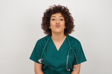 Pleasant looking young doctor female wearing medical uniform, keeps lips as going to kiss someone, has glad expression, grimace face. Standing indoors. Beauty concept.