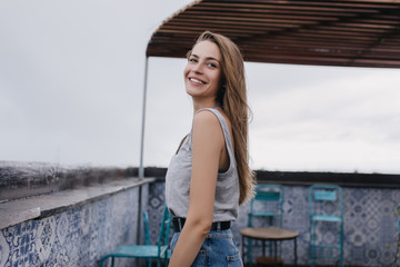 Slim positive girl standing at roof and posing with pleasure. Outdoor shot of attractive long-haired model standing on sky background with happy smile.