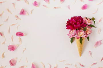 Creative flat lay with floral ice cream - fresh pink peonies in a waffle cone on a white background. Frame of pale pink petals, free space for text in the center. Mother's Day celebration concept.