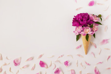 Spring creative flat lay with flower ice cream - lilac peonies in a waffle cone on a white background. Frame of pale pink petals, free space for text in the center. Mother's Day celebration concept.