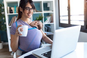 Pretty young mother with her baby in sling drinking coffee while working with laptop at home.