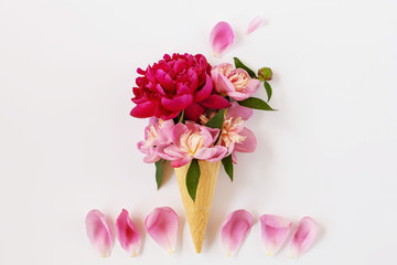 Creative floral still life of an ice cream waffle cone with peony flowers and pink petals on white surface. Beautiful botanical greeting card or postcard design, empty space for text. Top view.