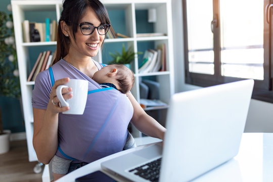 Pretty Young Mother With Her Baby In Sling Drinking Coffee While Working With Laptop At Home.