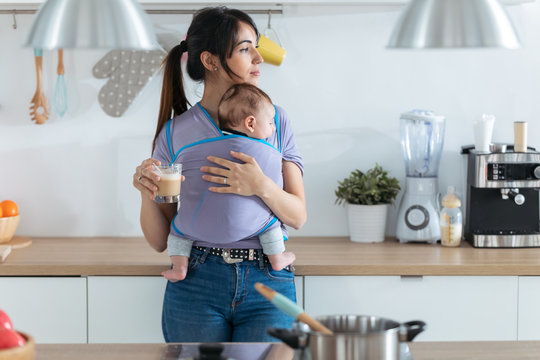 Pretty young mother with little baby in sling drinking coffee in the kitchen at home.