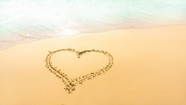 A heart shape written on golden brown sand beach beside pastel green color sea 's water