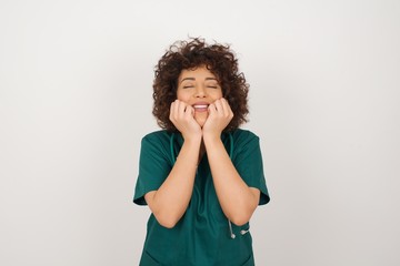 Portrait of young cute  doctor woman being overwhelmed with emotions, expressing excitement and happiness with closed eyes and hands near face while smiling broadly over gray background.