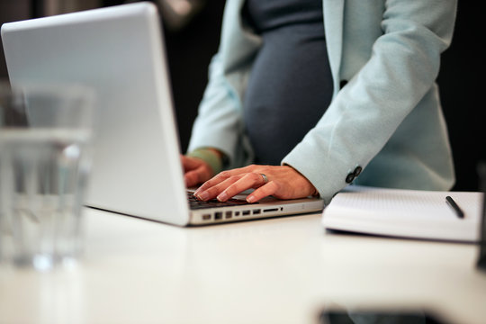 Close up of hardworking dedicated pregnant business woman standing in her office and typing report on laptop.	