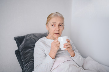 Coronavirus fever symptom senior woman drinking medicine or hot tea lying in bed at home quarantine. Corona virus infection causes respiratory illness first symptoms cough, headache, high temperature