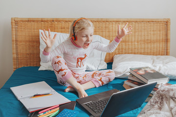 Caucasian girl child sitting in a bed and learning online on laptop Internet. Virtual class lesson...