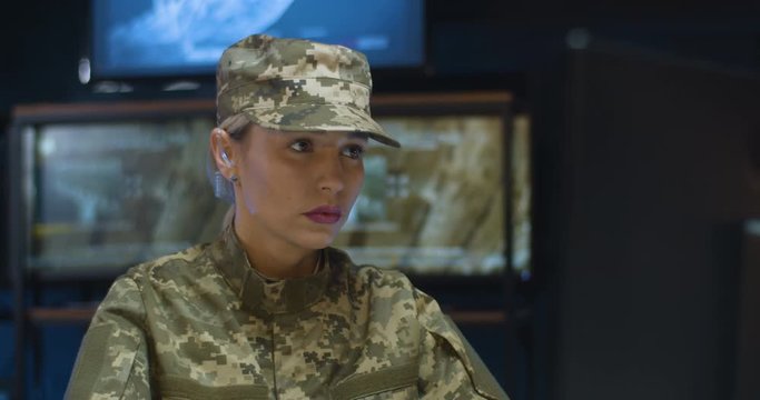 Close up of young pretty woman wearing camouflage clothes sitting at laptop computer and working in monitoring room in army. Female soldier in military data center. Beauiful lady works in troop.