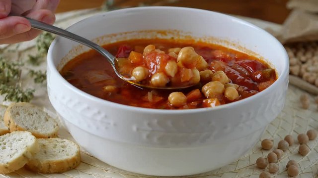 Healthy eating, take tablespoon chickpeas soup with tomatoes in a bowl. On the table a bowl of creamy and rustic legume soup. Eating healthy vegetarian dinner. Cozy and warm food. Protein dinner.