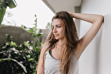 Fototapeta premium Brown-haired girl posing with dreamy face expression near white wall. Pensive caucasian lady touching her long hair and looking away.