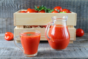 Fresh tomato juice in a glassware. A glass mug of tomato juice and fresh tomatoes in a wooden crate are on a gray wooden background. 
