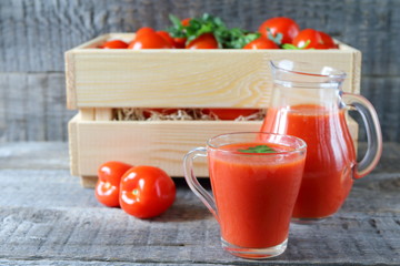 Fresh tomato juice in a glassware. A glass mug of tomato juice and fresh tomatoes in a wooden crate are on a gray wooden background. 
