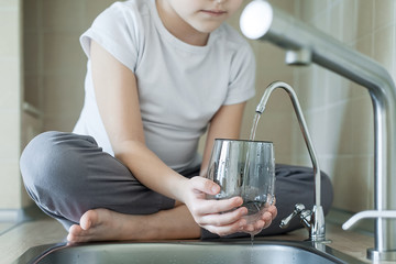 Child is using filter to purify drinking water. Kitchen faucet. Filling cup beverage. Pouring fresh...
