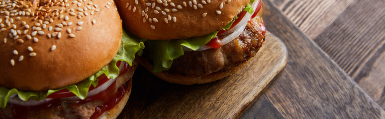 panoramic high angle shot of burgers on wooden cutting board, cutlery, oil, vinegar and beer bottles