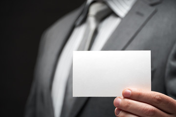 white blank business card closeup in businessman hand, gray suit, dark wall background