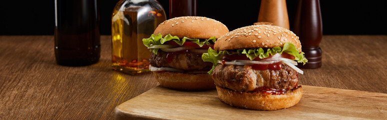panoramic shot of two tasty burgers on wooden board, cropped oil bottles, salt and pepper mills isolated on black