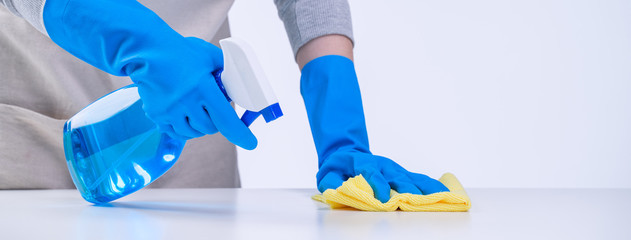 Young woman housekeeper in apron is cleaning, wiping down table surface with blue gloves, wet yellow rag, spraying bottle cleaner, closeup design concept.