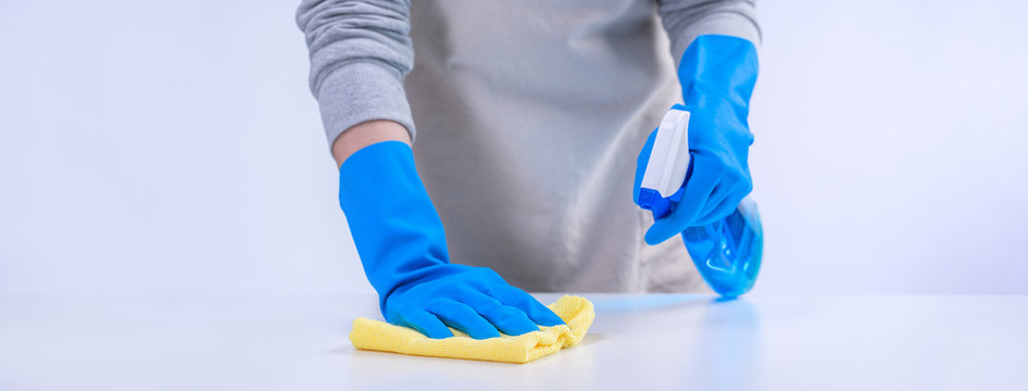 Young Woman Housekeeper In Apron Is Cleaning, Wiping Down Table Surface With Blue Gloves, Wet Yellow Rag, Spraying Bottle Cleaner, Closeup Design Concept.