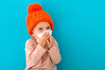 Child covers her face with her hands. Displeased little girl in red hat medical mask looking away on blue background