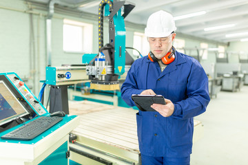 CNC machine operator in the Hard  white Hat Walks Through Light Modern Factory While Holding tablet. Successful, Handsome Man in Modern Industrial Environment.