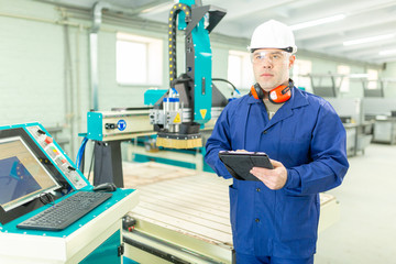 CNC machine operator in the Hard  white Hat Walks Through Light Modern Factory While Holding tablet. Successful, Handsome Man in Modern Industrial Environment.