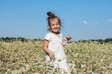 Happy joyful little girl in white linen dress standing on flowery meadow or field. Cute cheerful child playing among summer or spring flowers outdoors. Nature, rural landscape, childhood concept