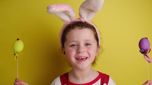Glad cheerful little girl covers eyes with two Easter eggs, wears bunny ears, dressed in white t-shirt, models over yellow wall with free space for promotion. Spring holiday and Easter concept.