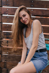 Enchanting european girl in short denim skirt posing on wooden background. Cute young lady with shy smile enjoying spring day.