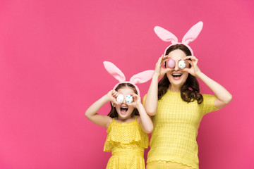 excited mother and daughter in bunny ears covering eyes with easter eggs isolated on pink