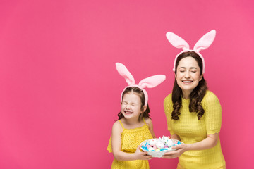 happy mother and daughter in bunny ears holding easter eggs and laughing isolated on pink