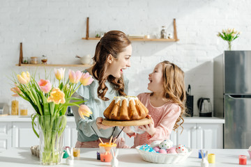 excited mother and child looking at each other near easter eggs, decorative rabbits, easter bread and tulips