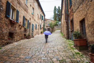 Fototapeta na wymiar Rear view of a girl with purple umbrella walking along a narrow picturesque medieval street of old town of Montepulciano in Tuscany, Italy